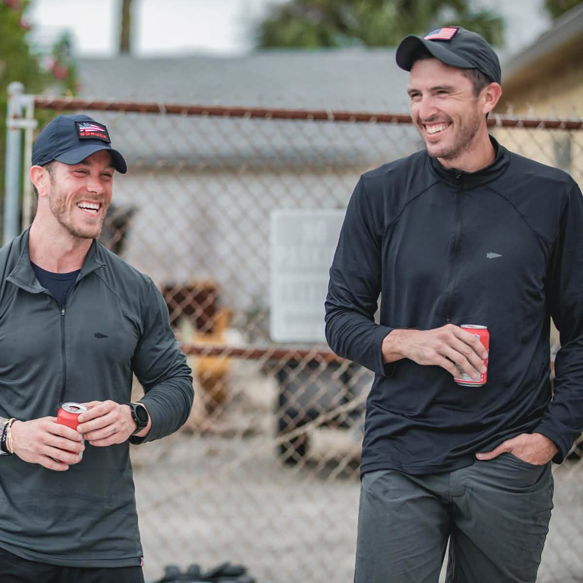 Two men in dark clothing and hats smile while holding drinks outdoors by a chain-link fence, each wearing The Indestructible Training Half Zip—premium workout apparel made from durable performance fabric for any occasion.
