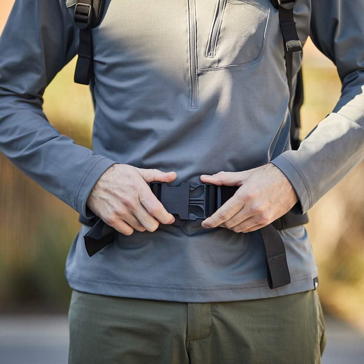 A person in a gray long-sleeve shirt and green pants expertly fastens the buckle on the black strap of their GORUCK backpack. The scene highlights their hands and the Padded Hip Belt, engineered for carrying heavy loads, set against a blurred outdoor background.