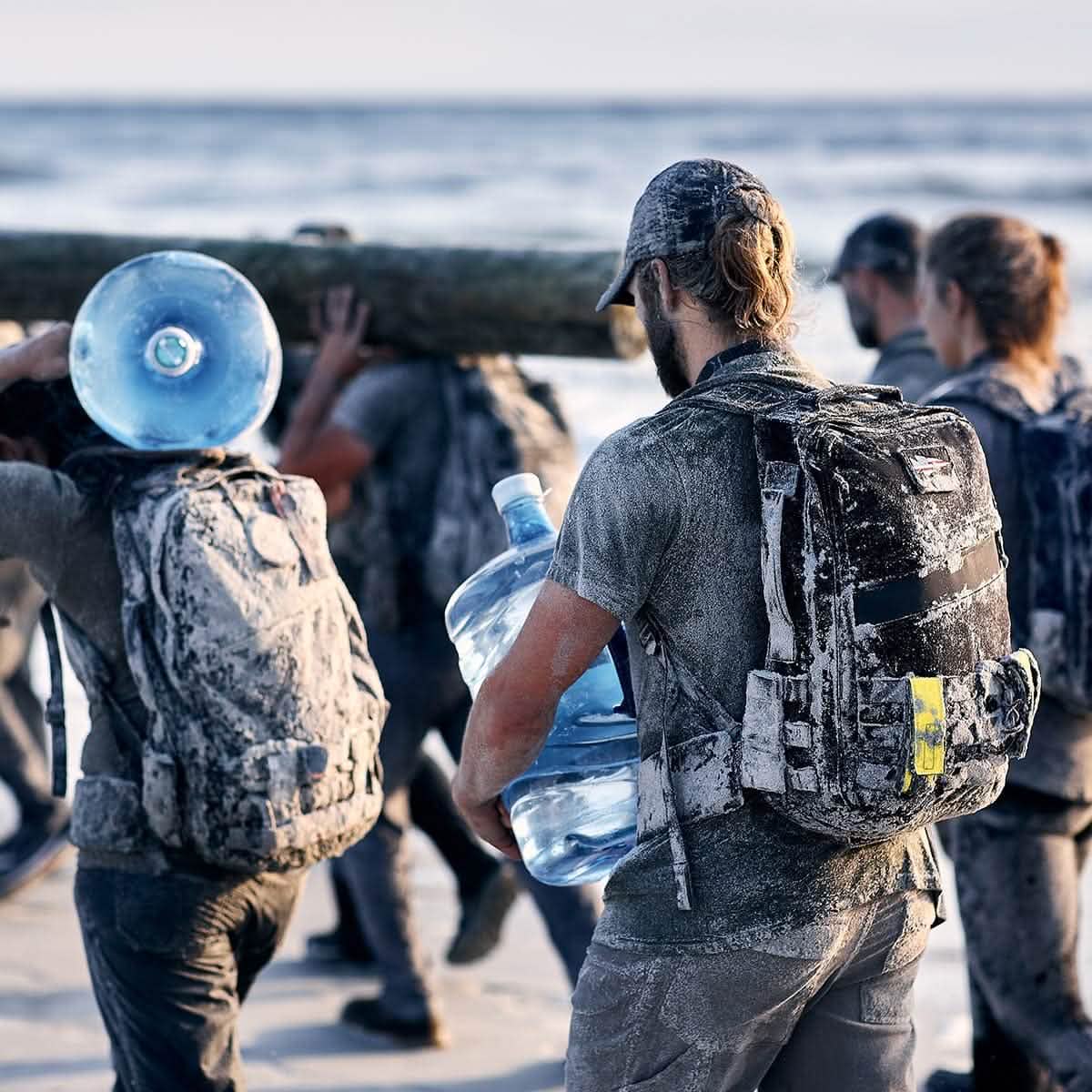 The group, dressed in outdoor gear and surrounded by sand, exercises intensely on the beach. One individual carries a large water jug secured with a GORUCK Padded Hip Belt, while others heft substantial loads such as a massive log. The scene is vibrant and implies teamwork and endurance in this coastal setting.