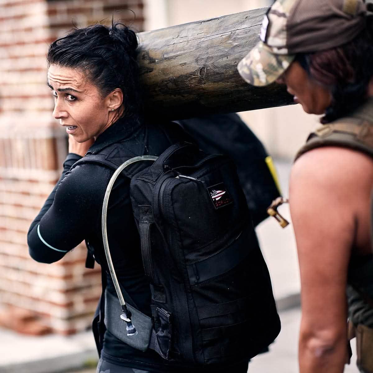 Two people carry a large wooden log on their shoulders. The woman in the foreground, equipped with a black vest and GORUCK backpack featuring a hydration tube, benefits from the comfort of a Padded Hip Belt. The person next to her wears a camo cap as they both appear focused and determined.