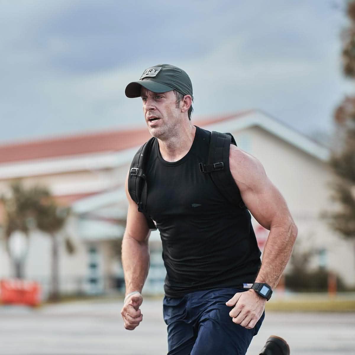Athletic man wearing a GORUCK Performance TAC hat, black tank top, and rucking backpack running outdoors near buildings
