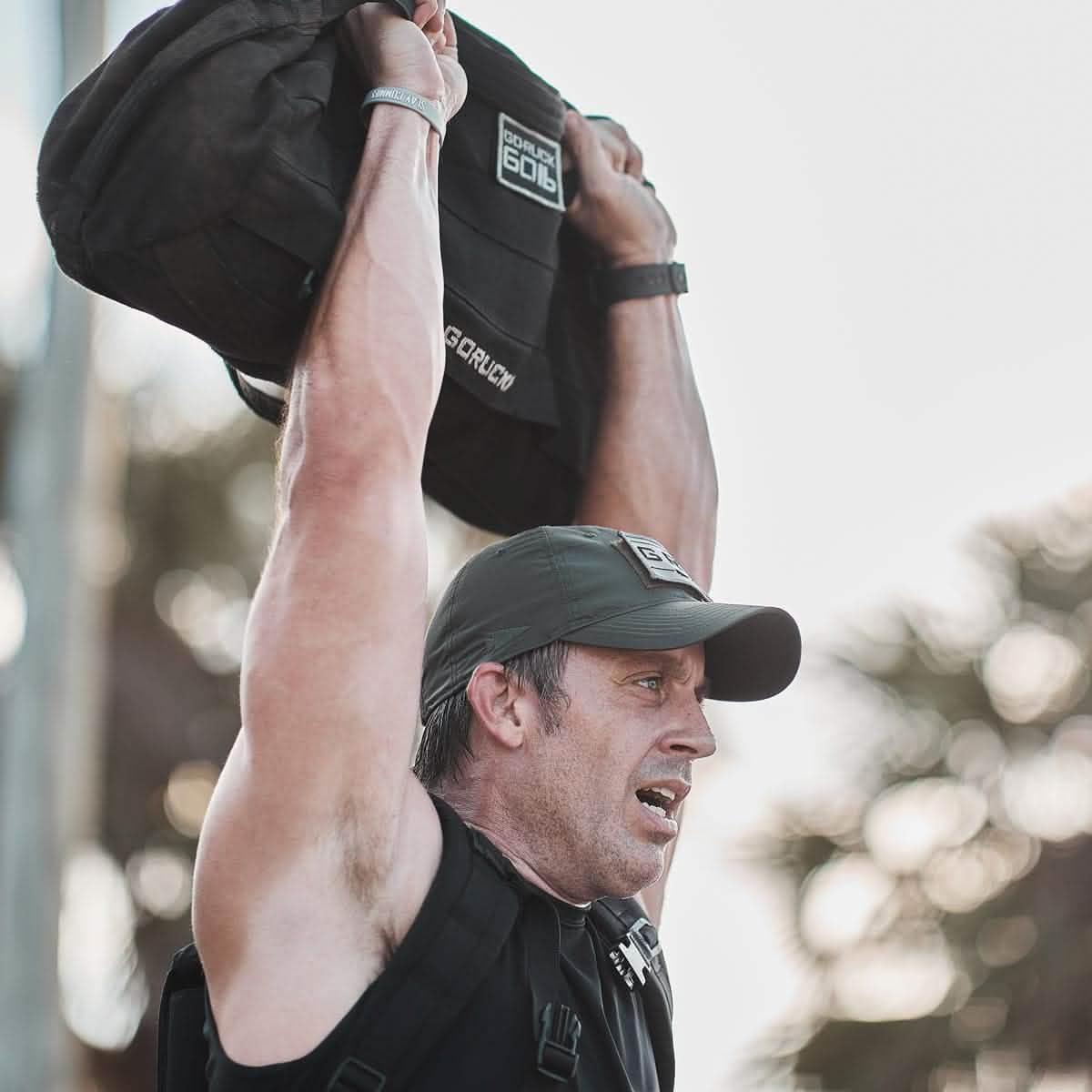 Man wearing GORUCK cap and vest lifting a GORUCK sandbag above his head during outdoor fitness training