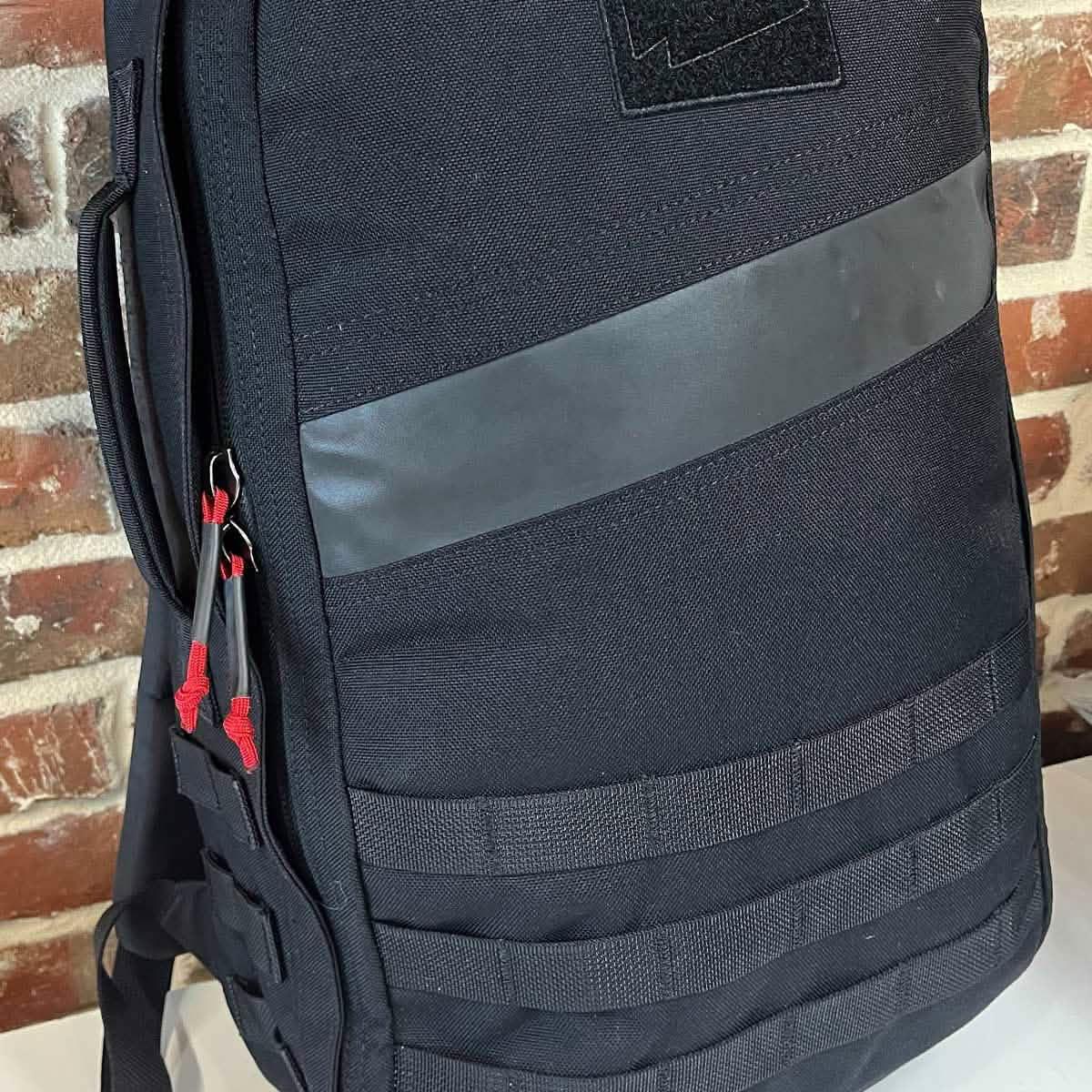 Close-up of black GORUCK rucksack with durable fabric, front MOLLE webbing, and red zipper pulls against brick wall
