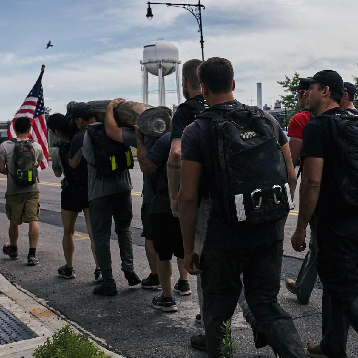 Group of people rucking with backpacks while carrying a log on a street near a water tower and American flag
