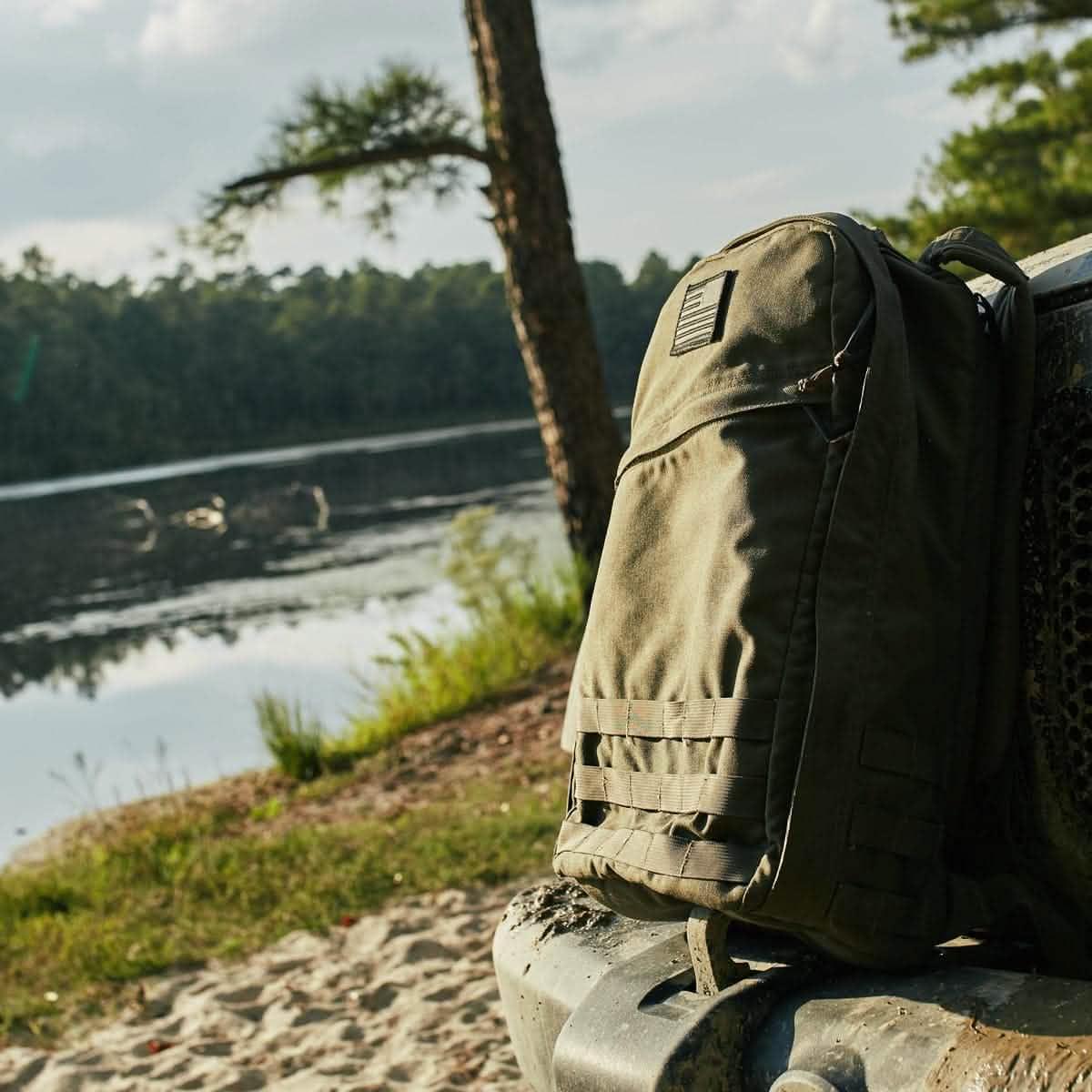 GORUCK olive green tactical backpack resting on a vehicle near a lakeside with trees and water in the background