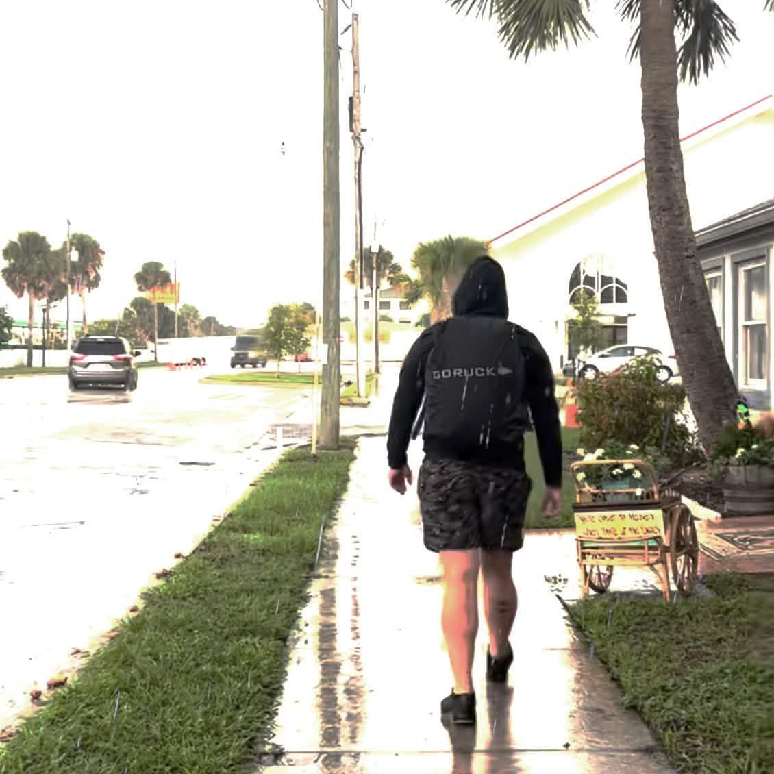 Person wearing black GORUCK backpack walking on wet sidewalk near palm trees and buildings