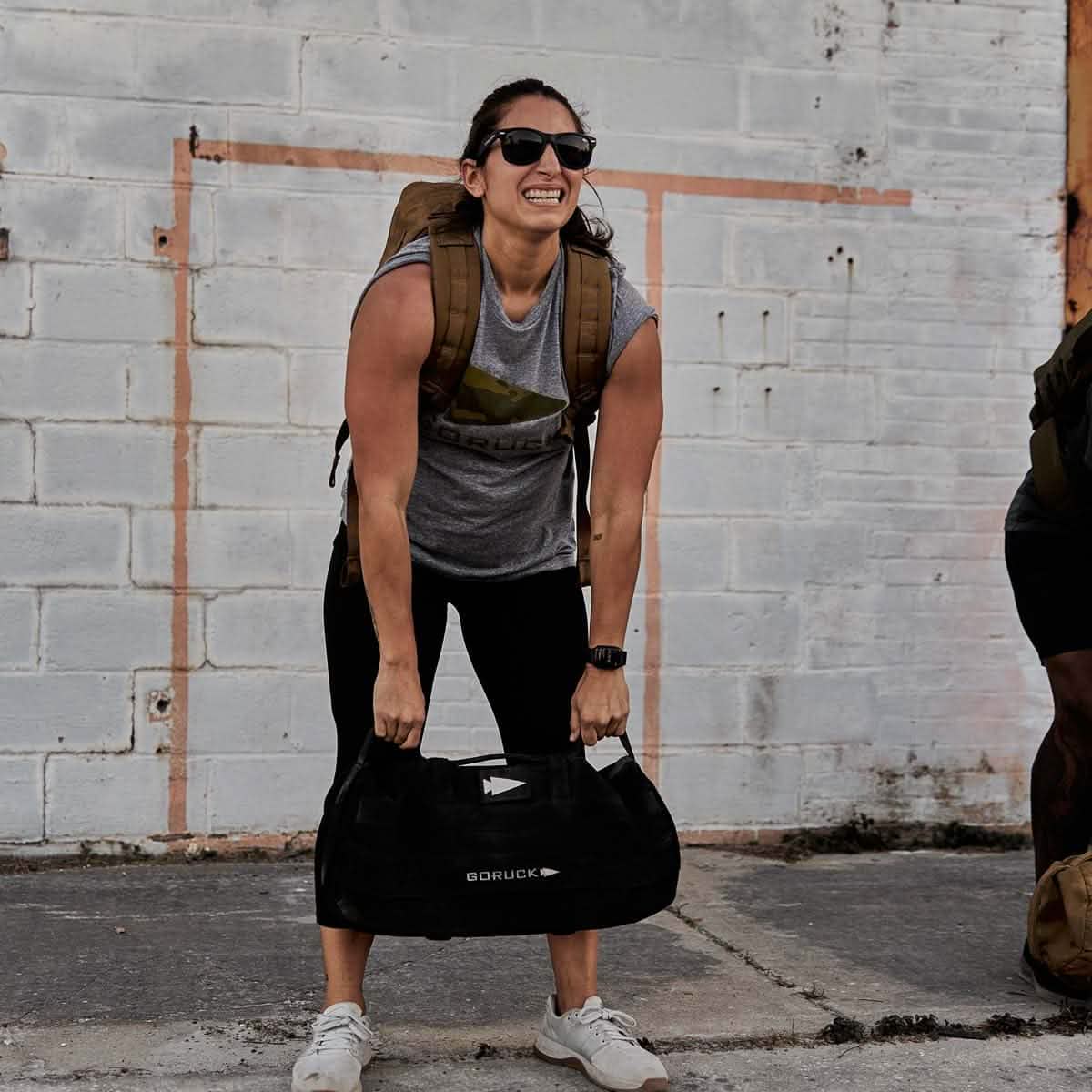 A person wearing sunglasses, a gray tank top, and black leggings lifts a GORUCK Sandbags 2.0 bag outdoors. Constructed from durable 1000D CORDURA, the bag appears ideal for their intense training routine in front of a white brick wall.