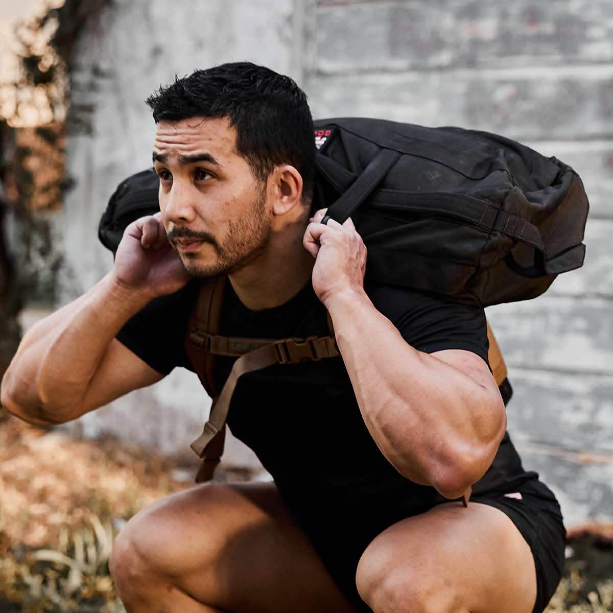 A man wearing a black shirt is squatting outdoors, carrying the GORUCK backpack made from robust 1000D CORDURA on his shoulders. The rustic building and grass indicate a natural setting, ideal for rugged adventures supported by the Scars Lifetime Guarantee of Sandbags 2.0.