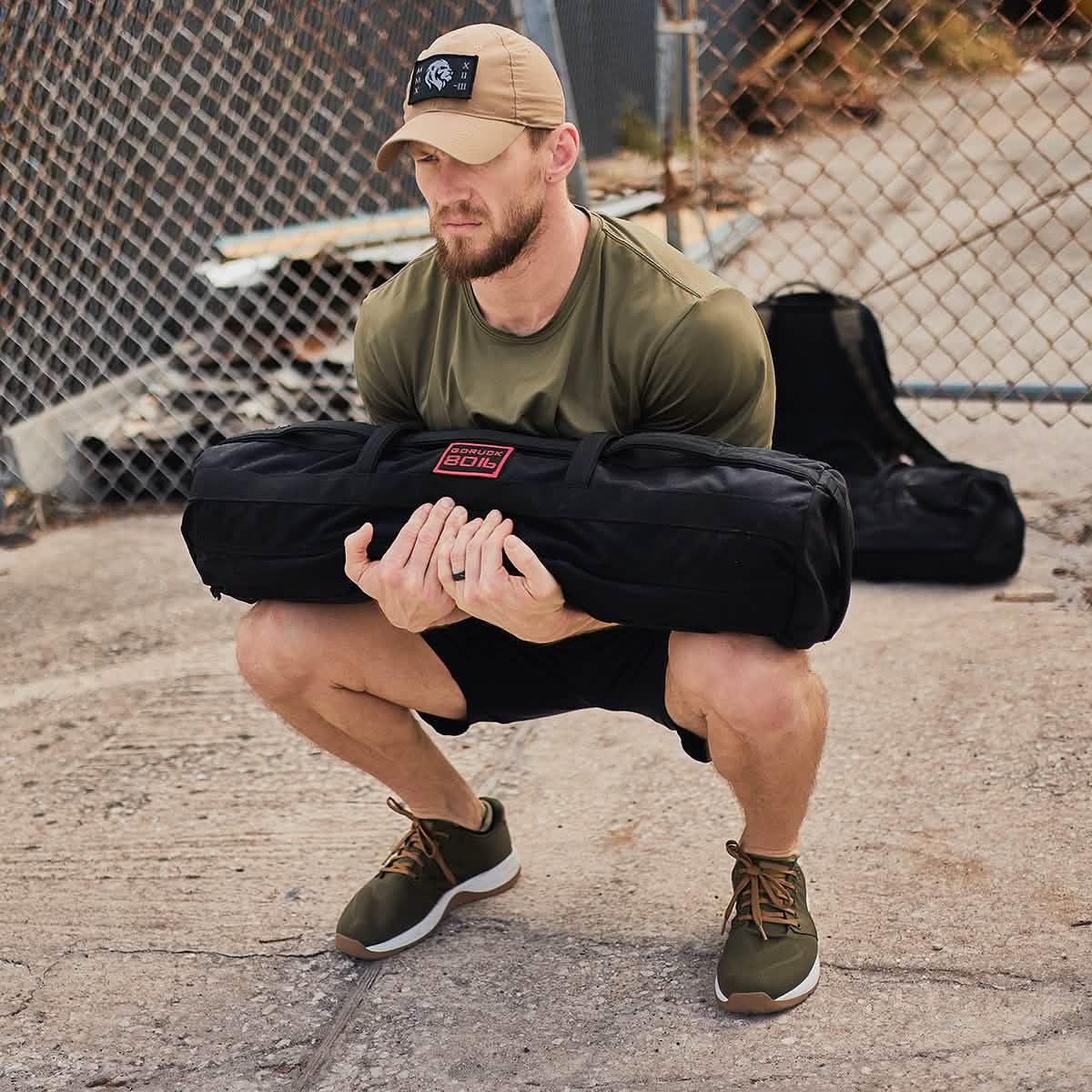 Man squatting with GORUCK training sandbag outdoors, wearing athletic gear