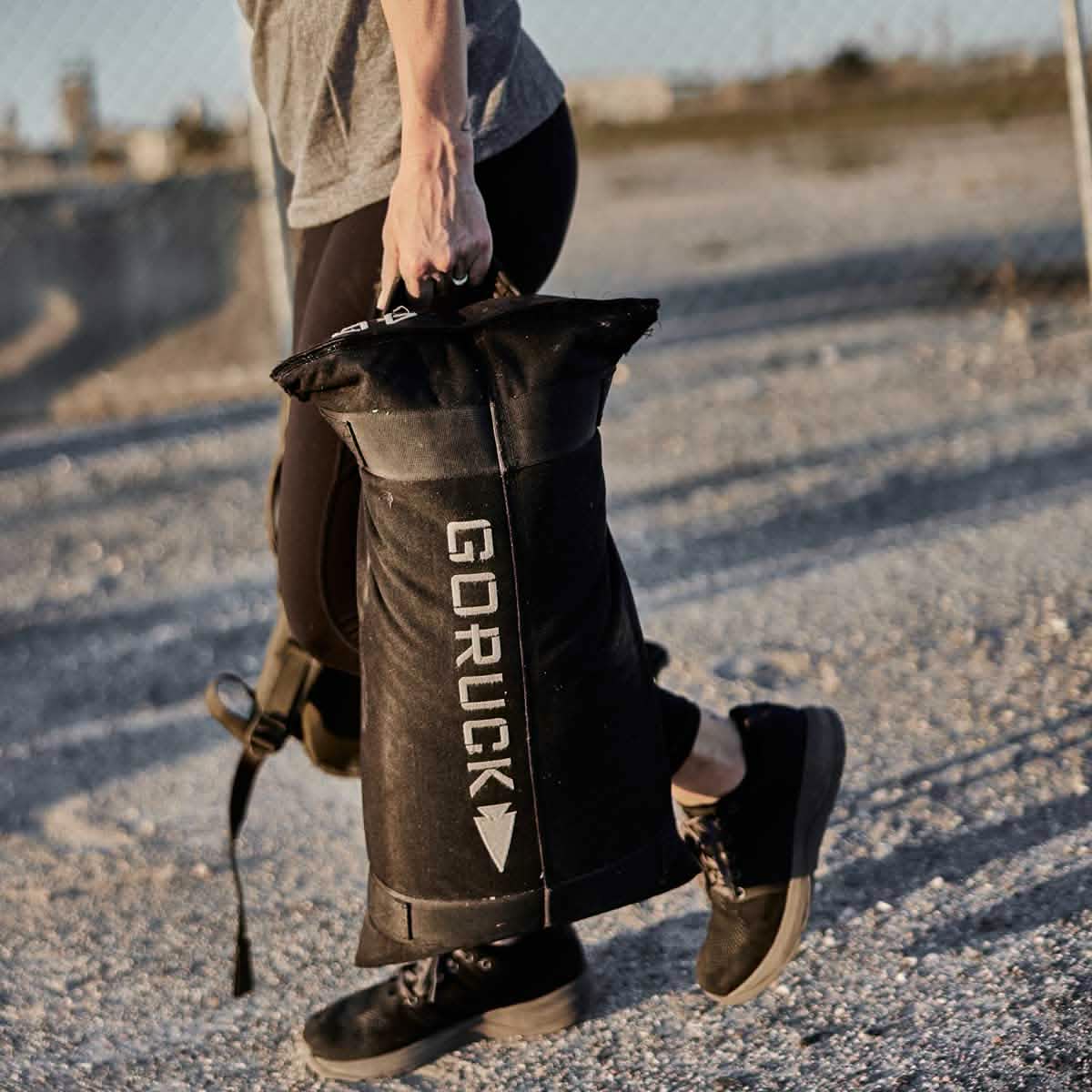 Dressed in athletic gear, an individual walks along a gravel path carrying a black Simple Training Sandbag by GORUCK. The long shadows cast by the sun accentuate their legs in black sneakers. In the distance, there's a chain-link fence against a blurred background, echoing the durability assured by GORUCK's Scars Lifetime Guarantee.