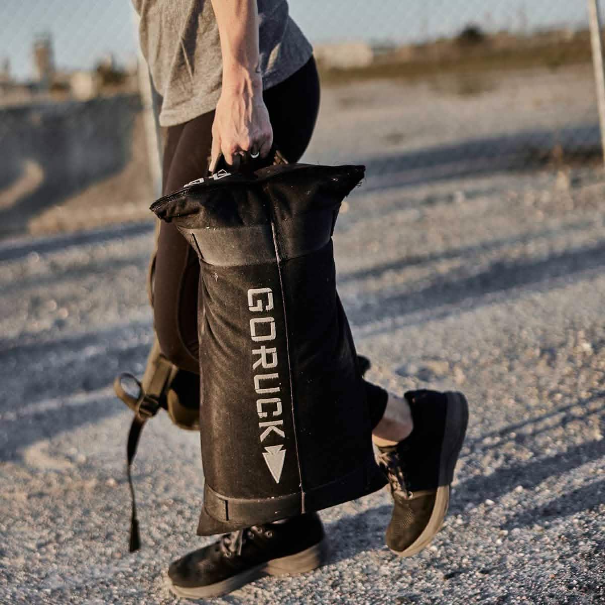 Person carrying a black GORUCK branded sandbag on a gravel outdoor surface for fitness training