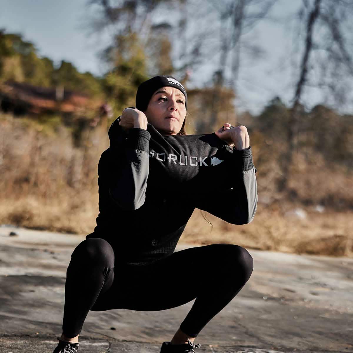 A person wearing athletic clothing and a beanie performs a squat outdoors on a sunny day, using GORUCK's Simple Training Sandbags for resistance, with trees and dry grass in the background.
