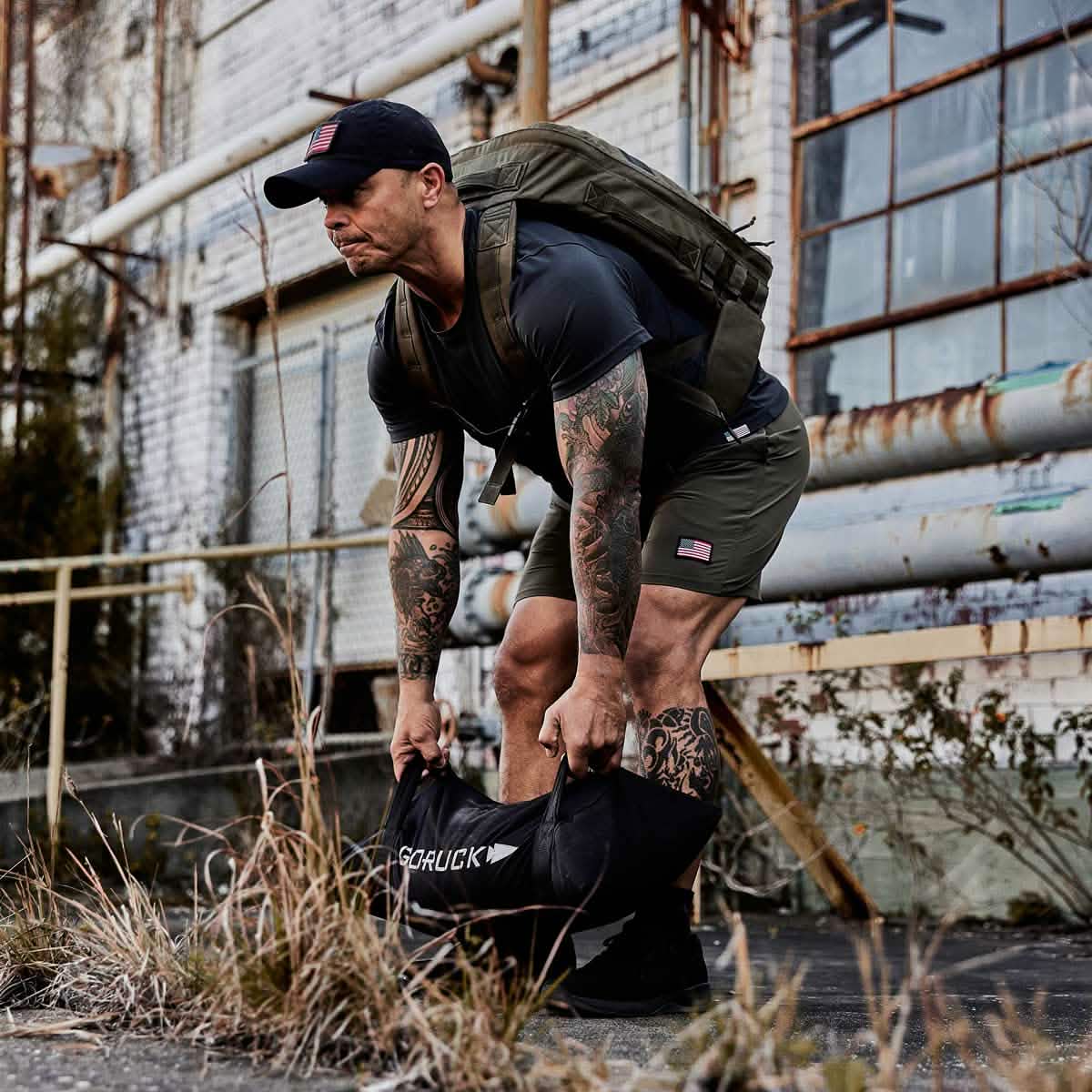 A man wearing a dark cap and shorts, showcasing tattoos on his arms and legs, is training with a black GORUCK Simple Training Sandbag outdoors near an industrial building. Carrying a backpack, he embodies the grit of outdoor workouts, counting on the Scars Lifetime Guarantee as he moves through the grass.