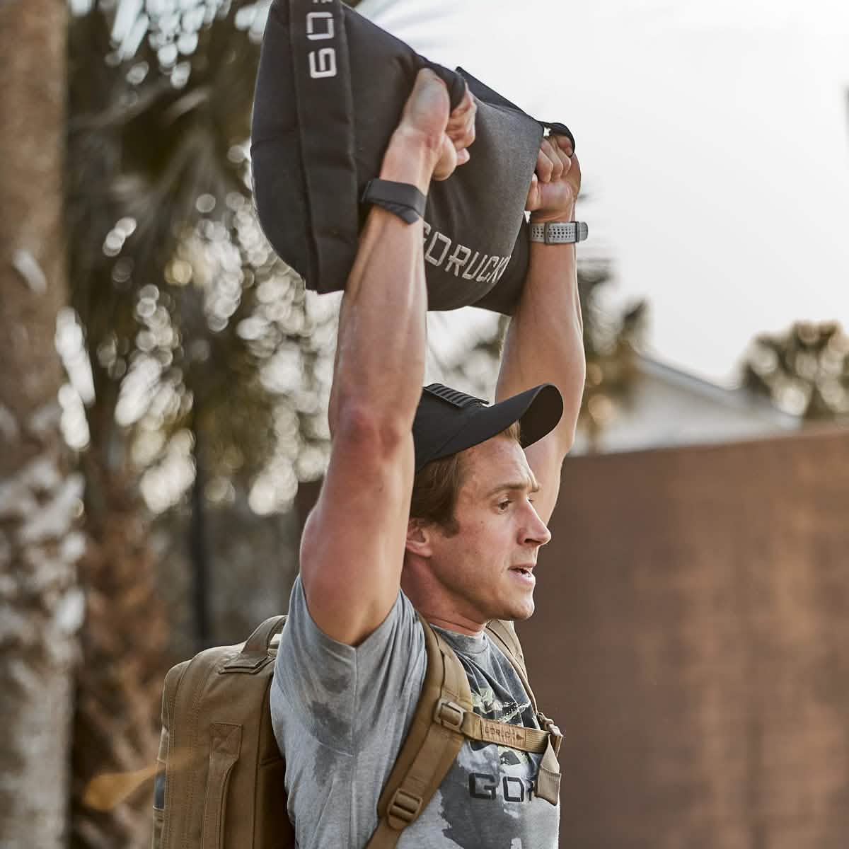 A person outdoors hoists a Simple Training Sandbag by GORUCK, featuring padded handles, overhead. Dressed in athletic gear and wearing a cap and backpack, they are framed by a building and palm trees in the background, all with the assurance of GORUCK's Scars Lifetime Guarantee.