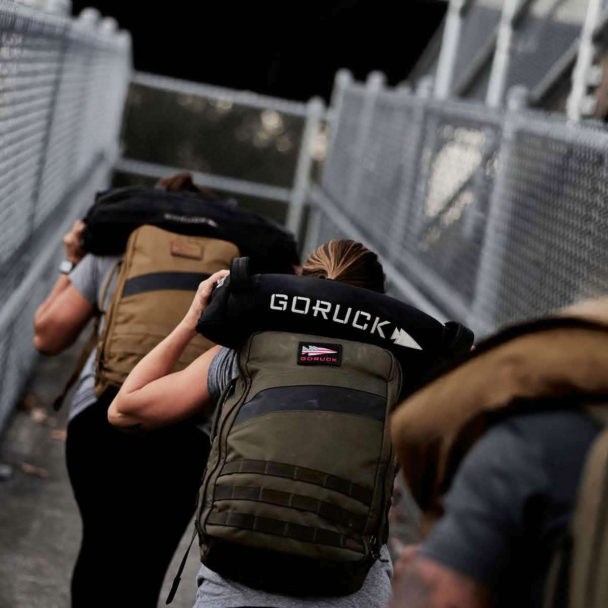 People wearing backpacks walk in a line on a fenced path, highlighting GORUCK's famous Scars Lifetime Guarantee. One participant carries a Simple Training Sandbag labeled GR1 USA, suggesting an intense outdoor fitness or rucksack training event.