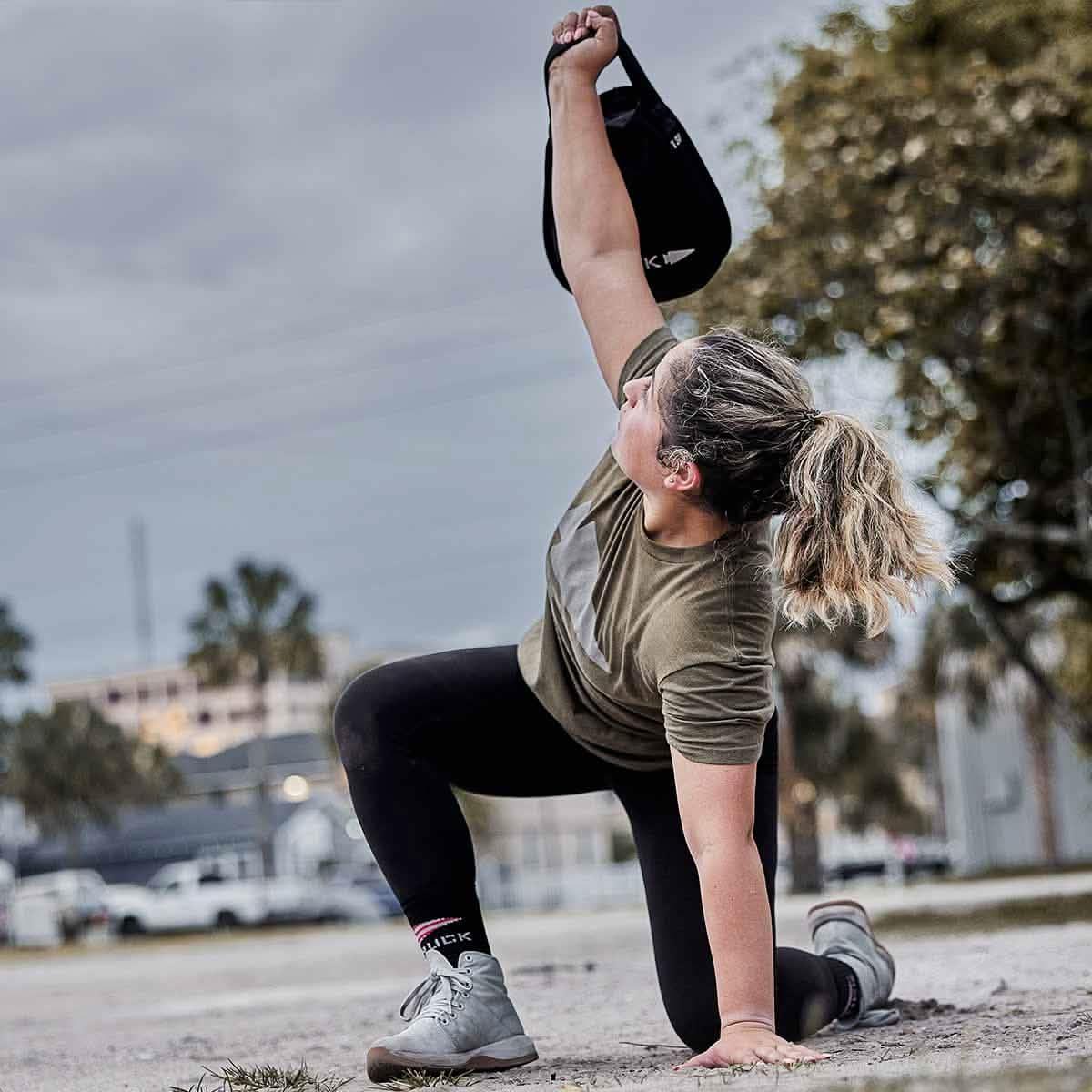 Person in athletic gear doing a dynamic stretch outdoors holding a black GORUCK sandbag