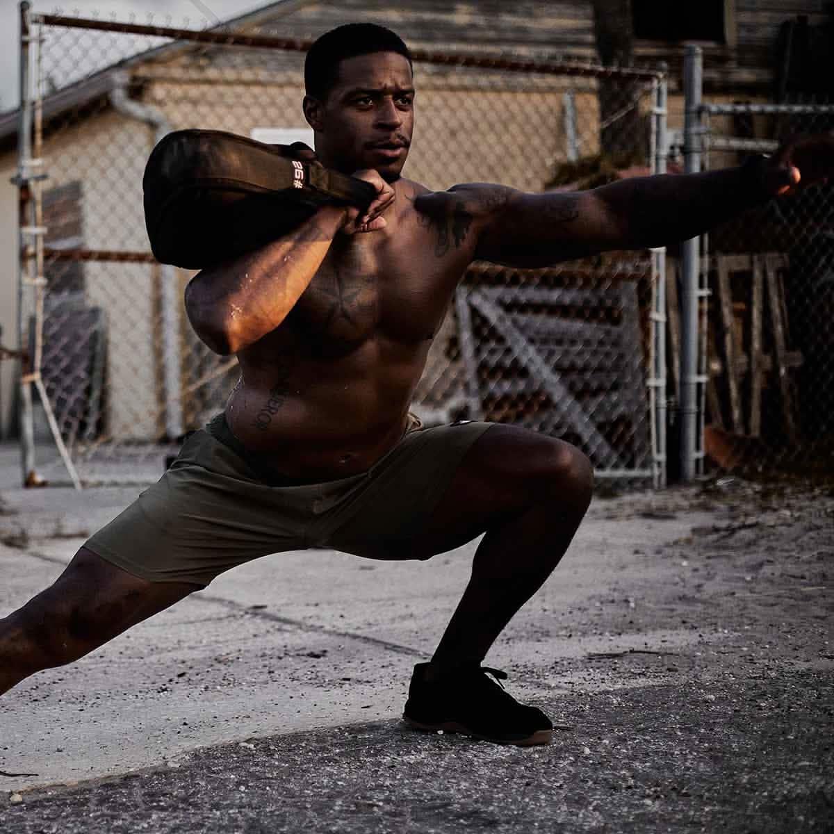 Shirtless muscular man performing side lunge exercise outdoors holding a GORUCK sandbag on shoulder
