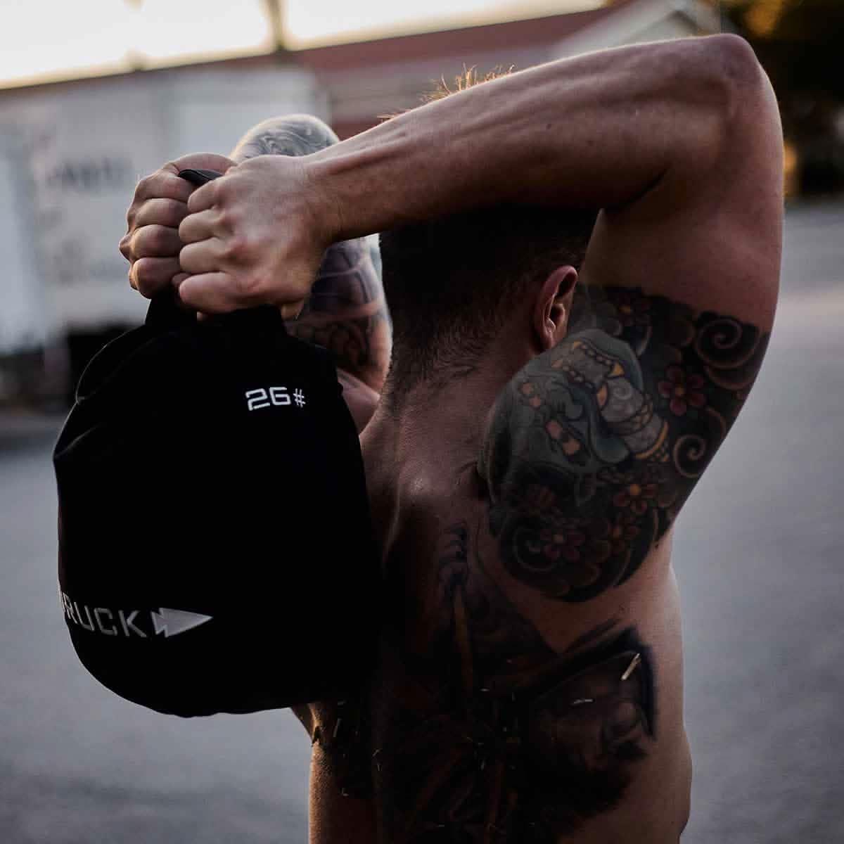 Muscular tattooed man holding GORUCK 26# weighted sandbag over shoulders outdoors