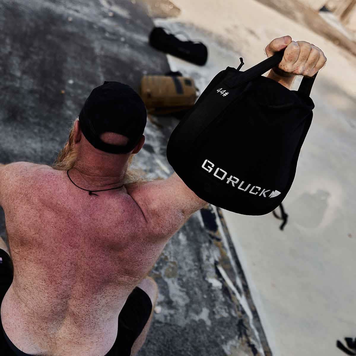 A sunburned individual, without a shirt, is using one arm to lift a GORUCK Sand Kettlebell and is donning a black cap while seated on a textured surface that highlights its endurance design. In the background, two sandbags can be seen.