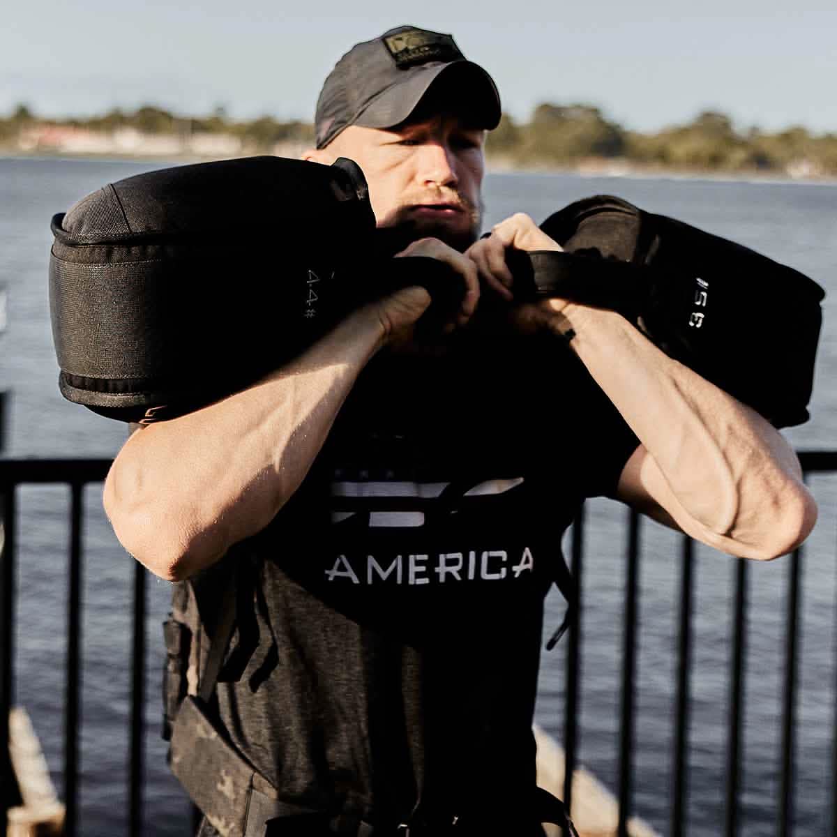 A person wearing a black cap and T-shirt is exercising with GORUCK Sand Kettlebells by a waterfront. Their arms are bent, holding the weights close to their shoulders. A metal railing and water are visible in the background, showcasing an endurance design perfect for fitness enthusiasts.