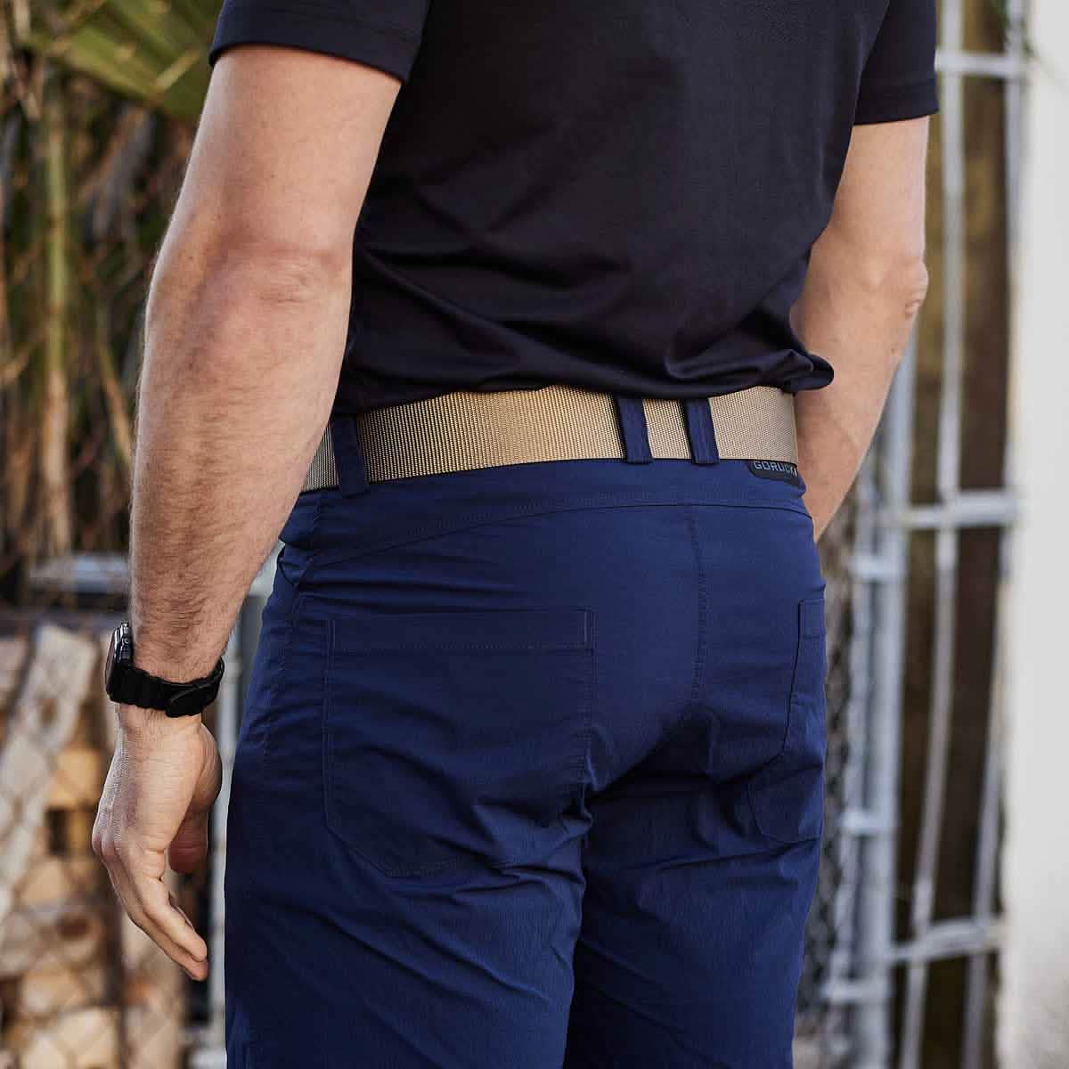 A man dressed in a navy blue shirt and pants along with a Grip 6 Spearhead Tactical Belt stands outdoors. On his left wrist, he's wearing a watch, while the background highlights greenery and a metal structure.