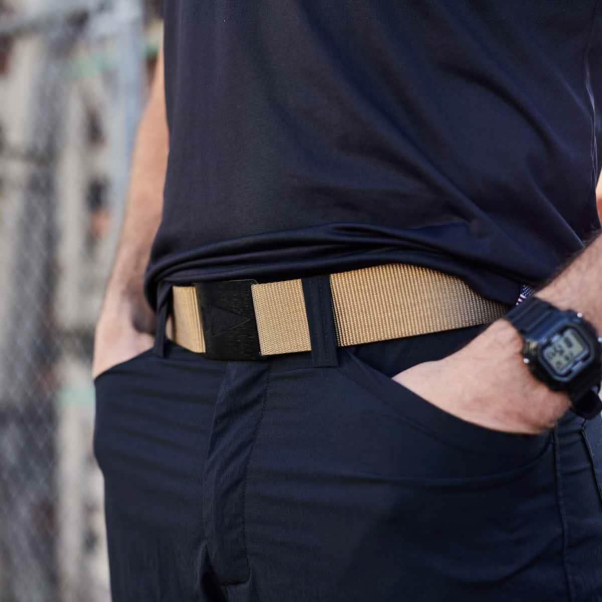 Close-up of a person wearing a tan tactical belt with a sturdy black buckle and navy pants, hands in pockets