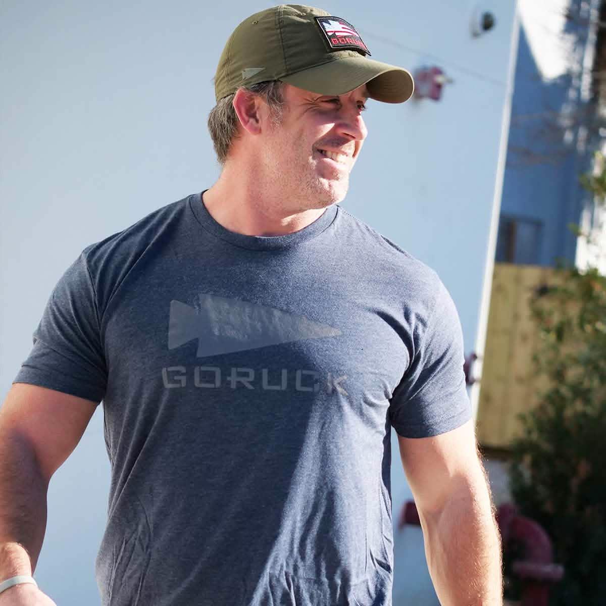 Man wearing navy blue GORUCK logo t-shirt and olive green cap outdoors in bright daylight