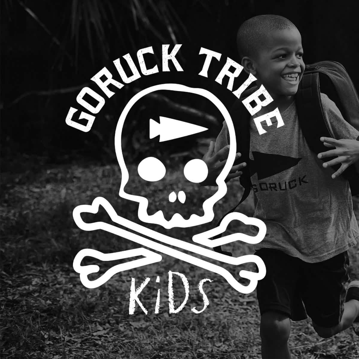A joyful child wearing a GORUCK shirt runs outdoors with a backpack, sporting the GORUCK TRIBE KIDS logo on display. The logo, featuring a skull, crossbones, and an arrow, symbolizes the adventure that awaits every young subscriber of the GORUCK Tribe Kids Monthly Subscription.