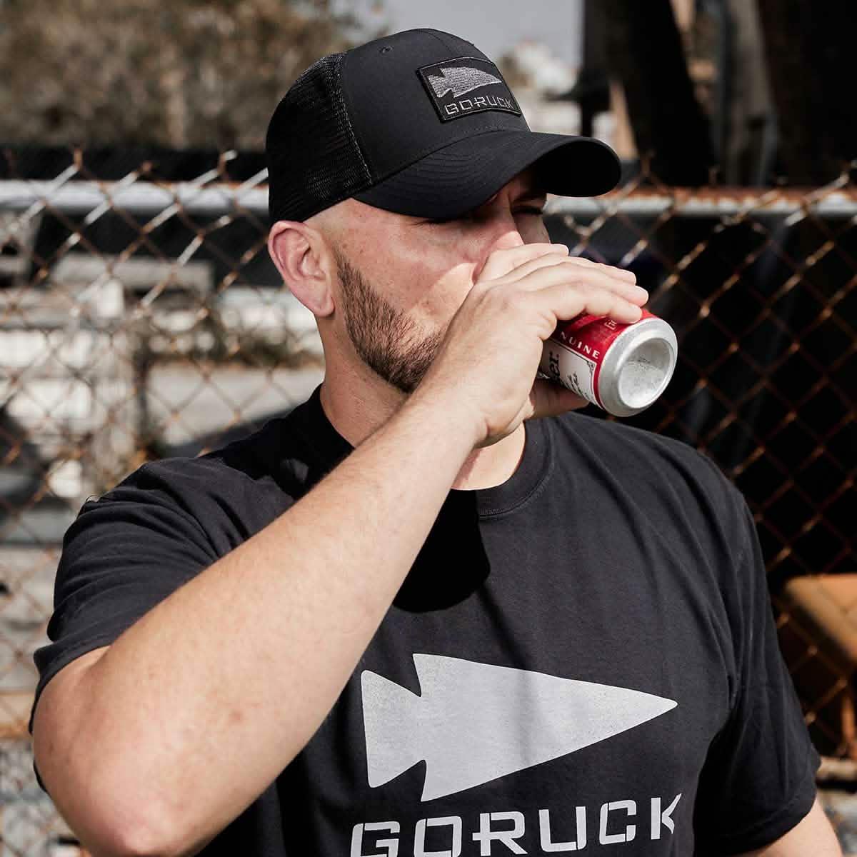 Man wearing black GORUCK hat and t-shirt drinking from a can outdoors by a chain-link fence.