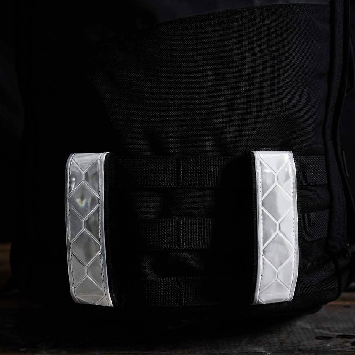 A close-up of a black bag showcases GORUCK's Reflective Ruck Bands, featuring a diamond pattern. The MOLLE webbing enhances its utility, and the shiny, textured material stands out against the dark background, ensuring visibility at night.
