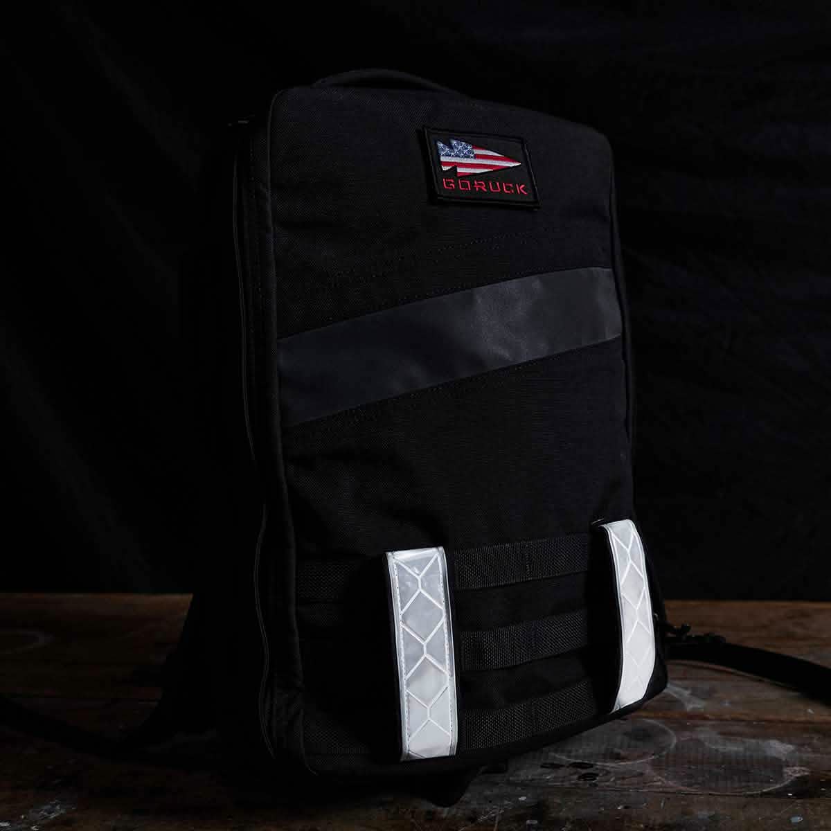 A black backpack featuring a GORUCK American flag patch on the front, enhanced with Reflective Ruck Bands including a horizontal dark green stripe and vertical white MOLLE webbing straps for improved utility. The backpack is set on a wooden surface, providing excellent nighttime visibility against a dark background.