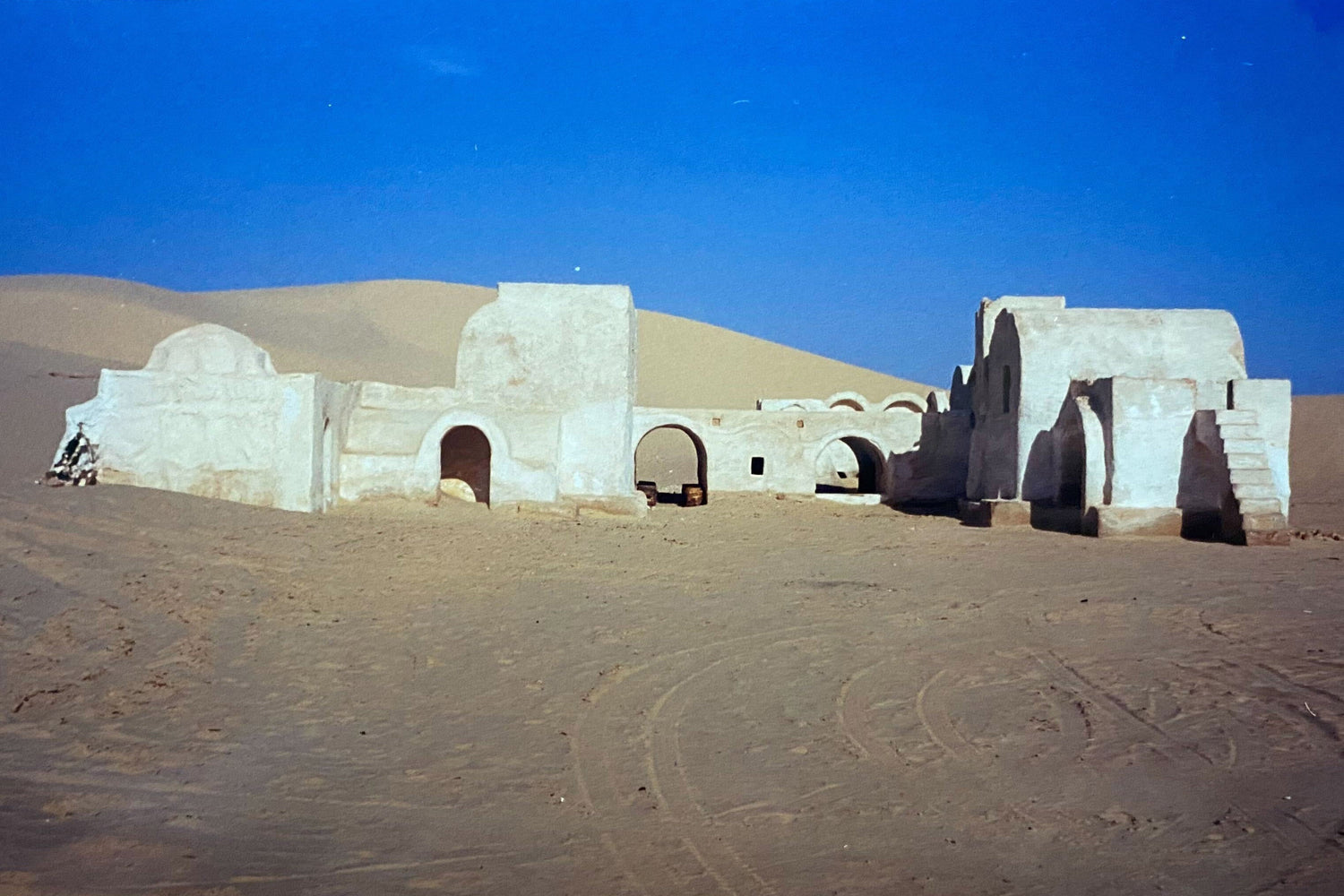 White adobe-style desert buildings with arches and stairs against sand dunes under clear blue sky