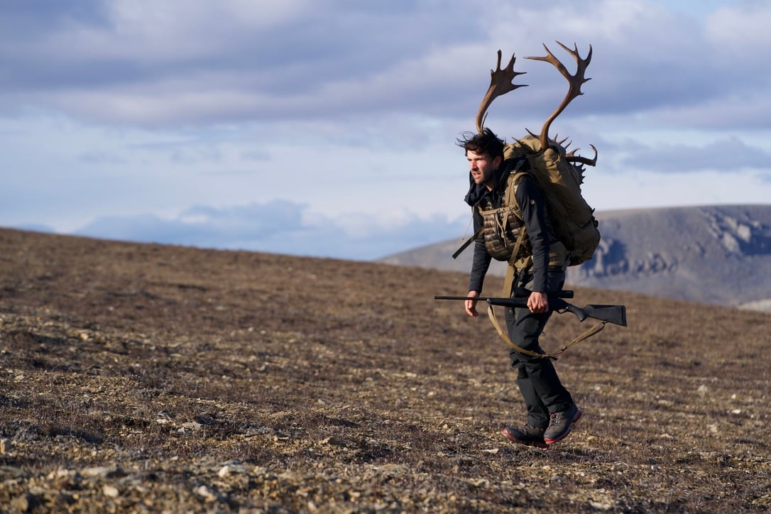 Man rucking outdoors with antlers and rifle, wearing GORUCK tactical backpack on rugged terrain