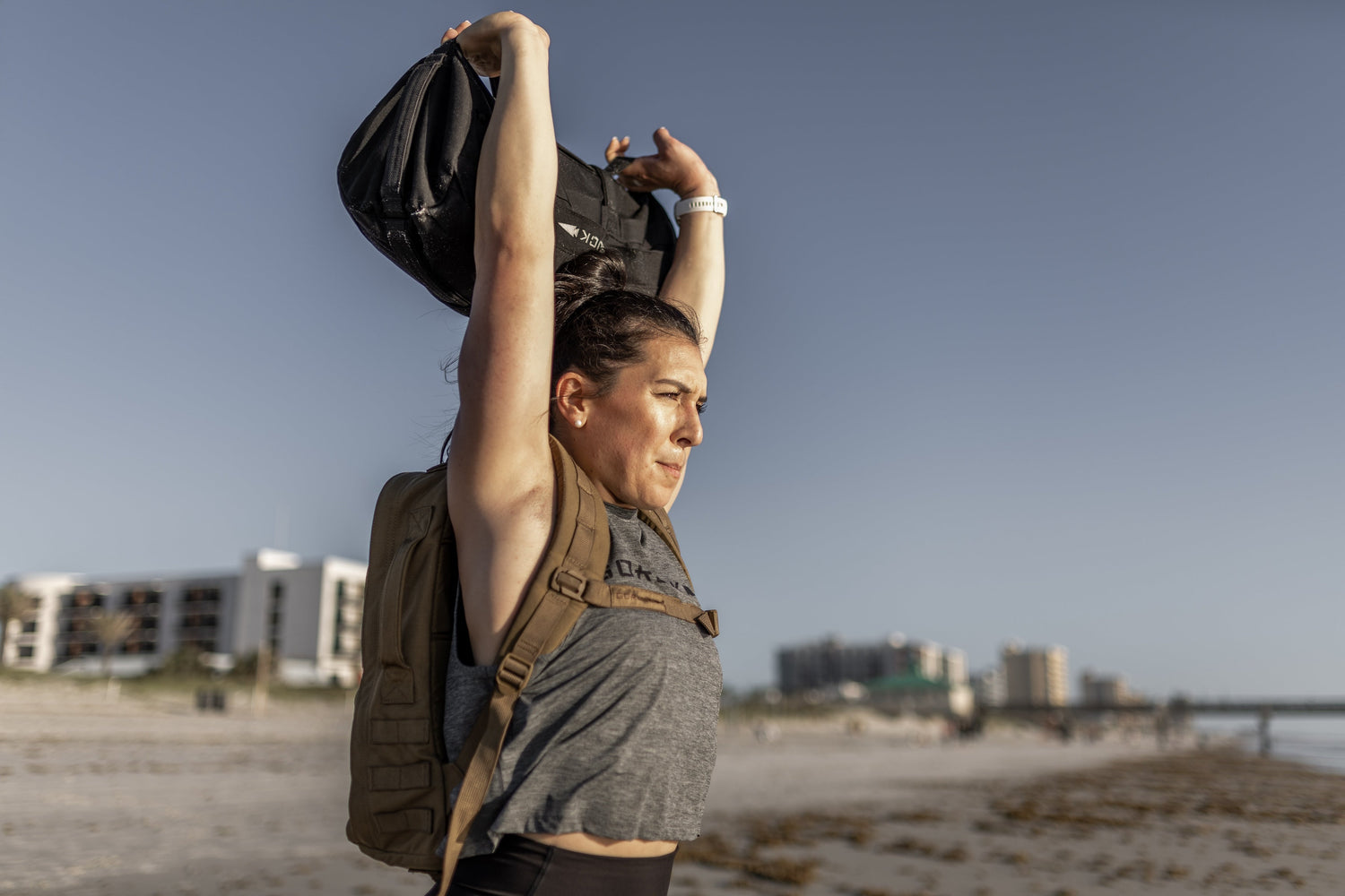Woman rucking on beach with GORUCK backpack and sandbag, buildings in background