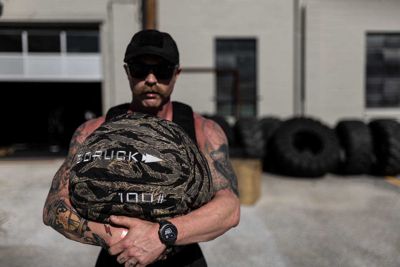Tattooed man outdoors holding GORUCK 100 lb training sandbag, large tires in background