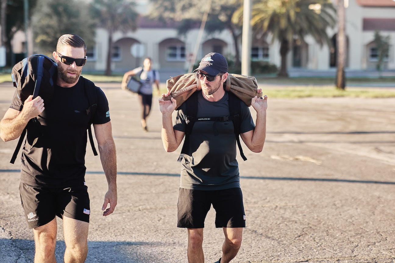 Two men rucking outdoors with GORUCK gear and sandbags, wearing athletic apparel