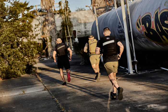 Three men rucking in tactical vests, running outdoors on concrete at GORUCK training event.