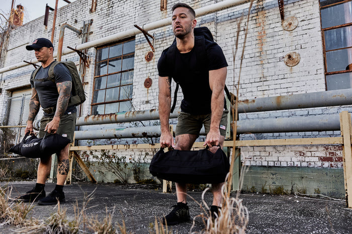 Two men rucking outdoors with GORUCK gear bags and backpacks in front of an industrial brick wall