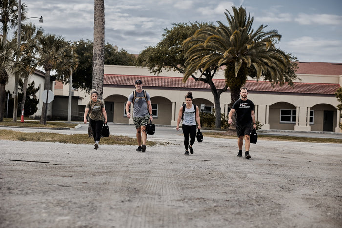 Four people rucking with GORUCK sandbags outdoors, palm trees and building in background