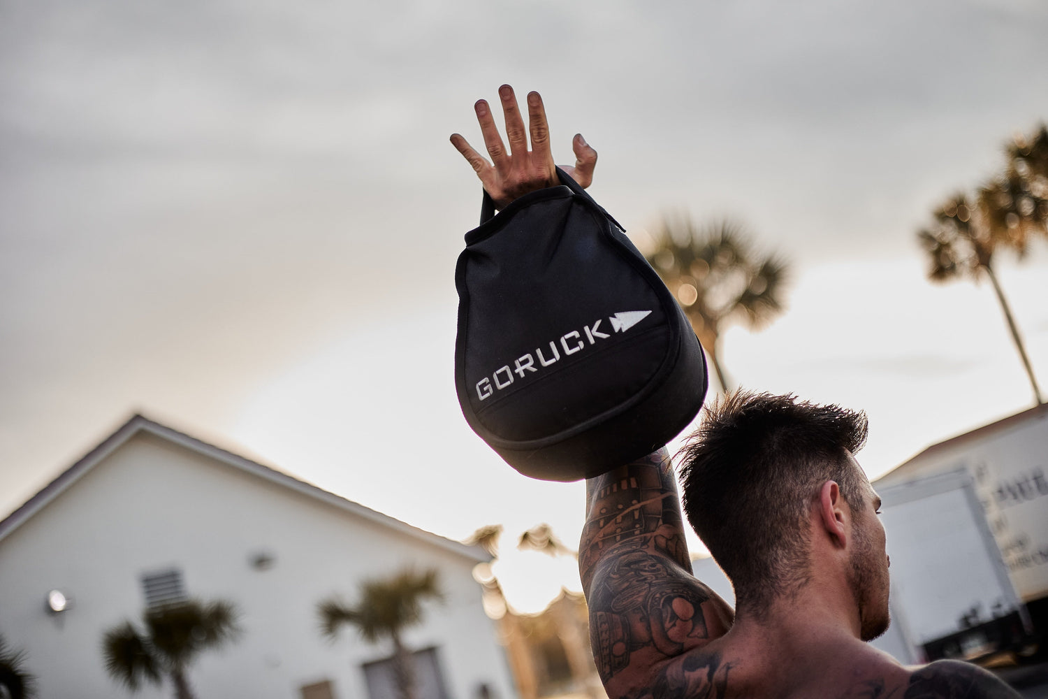Tattooed man lifting GORUCK sandbag outdoors near palm trees at sunset