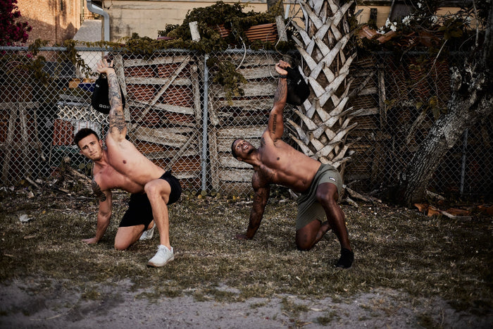Two athletic men perform outdoor sandbag exercises with GORUCK gear near a fence and palm tree.
