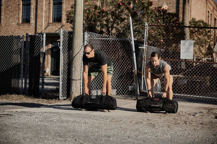 Two people lifting GORUCK sandbags outdoors, training with rucking gear near a fence.
