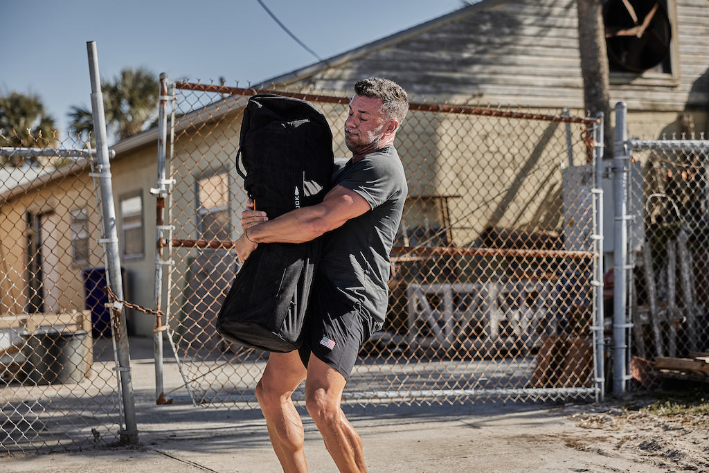 Man outdoors lifting GORUCK sandbag, showcasing rugged rucking gear in urban setting