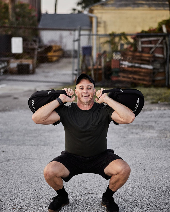 Man doing outdoor squat with GORUCK sandbag, wearing black athletic gear, urban background