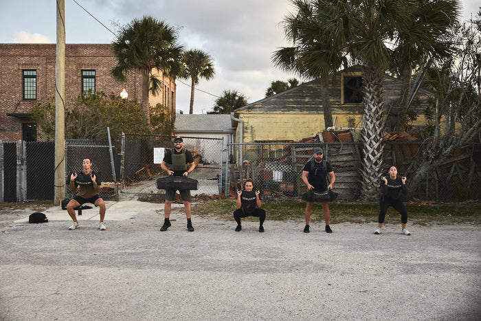 Five people doing outdoor rucking exercises in GORUCK gear, brick buildings and palm trees behind.