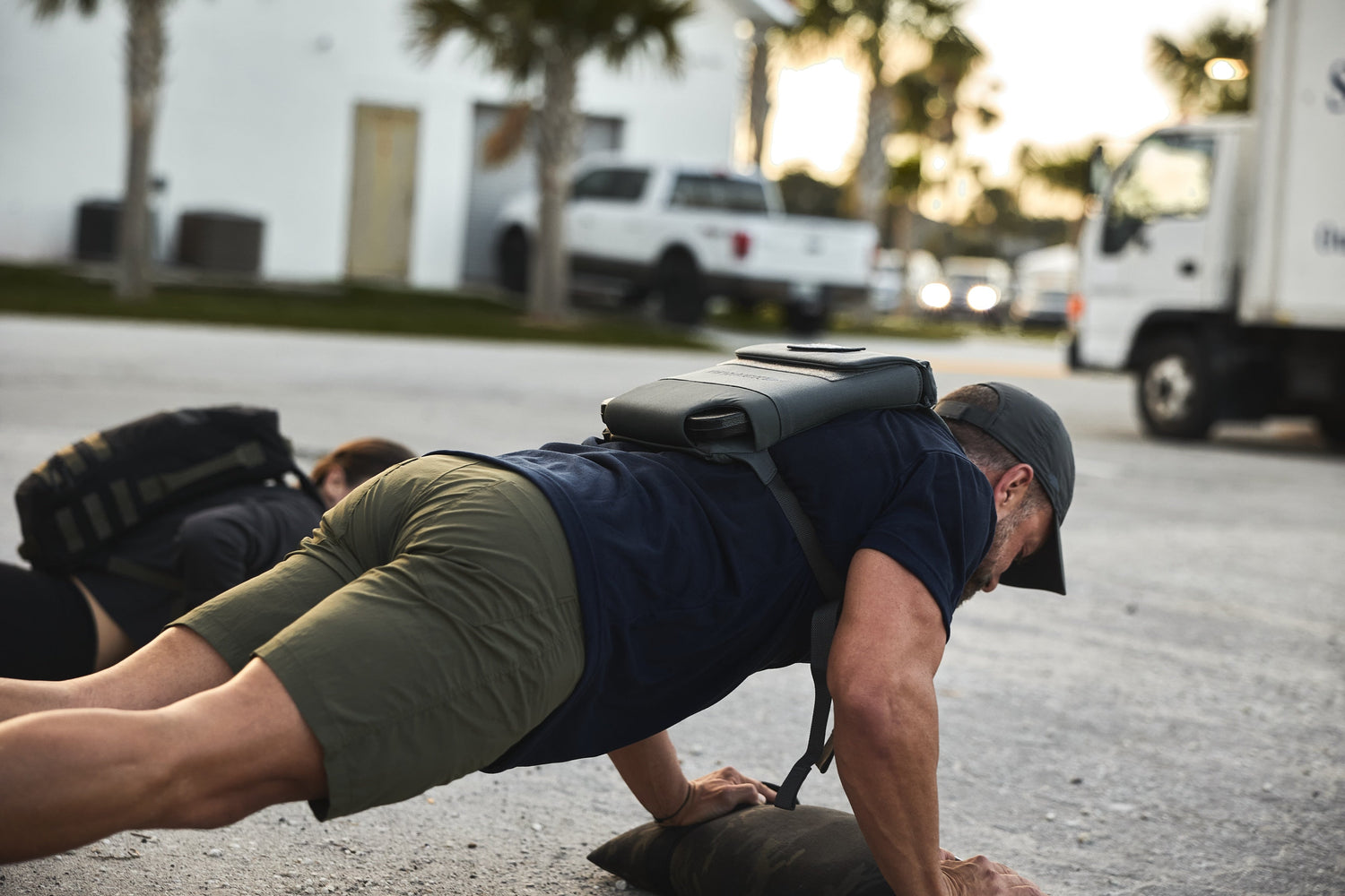 Man doing outdoor push-ups wearing GORUCK training backpack and cap on pavement