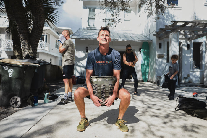 Man squatting outdoors with GORUCK sandbag, group training in sunlight, fitness gear visible