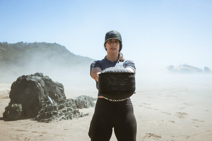 Woman training with GORUCK sandbag on a sandy beach, wearing GORUCK cap and athletic gear.
