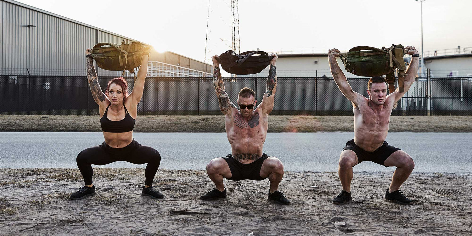 Three fit individuals performing overhead squats with weighted backpacks outdoors on sandy ground