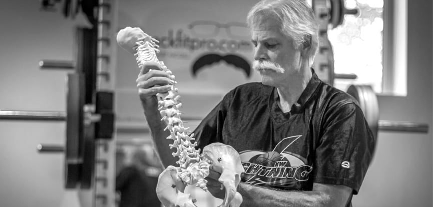 Older man examines anatomical spine model in gym setting with weights in background
